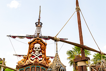 The image showcases the stern of a pirate ship with a skull and crossbones emblem, rigging, and mast against a pale blue sky at Disneyland Paris.