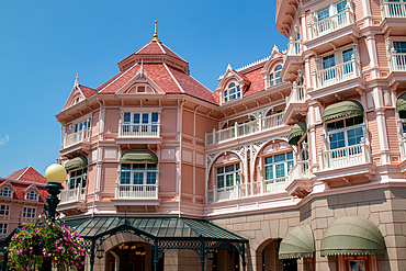 Facade of a pink building with white trim, red tile roof, green awnings, and balconies under a clear blue sky at Disneyland Paris.