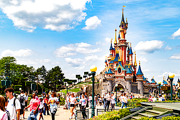 Crowd of people in front of a fairytale castle under a bright blue sky with fluffy white clouds on a sunny day at Disneyland Paris.