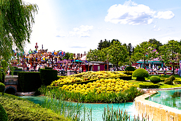 A vibrant park scene with water features, manicured hedges, a yellow flower bed, a carousel ride, and a crowd of people under a blue sky at Disneyland Paris.