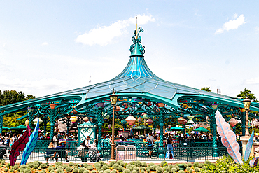 A teal-colored, ornate carousel shelter is filled with people and spinning teacups, beneath a blue sky with scattered clouds at Disneyland Paris.