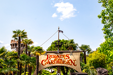 A shot showcasing the 'Pirates of the Caribbean' entrance sign, framed by palm trees, greenery, and a turret against a bright blue sky at Disneyland Paris.