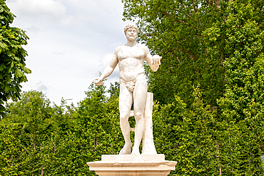 Full shot of a nude male statue on a pedestal holding fruit, surrounded by lush green foliage under a partly cloudy sky at Palace of Versailles, Paris.