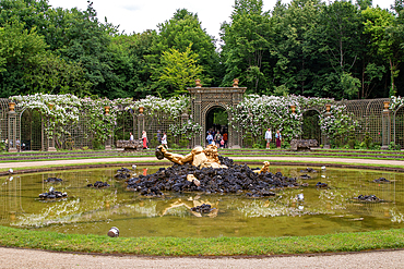 A golden statue lies partially submerged in a pond in a formal garden with trellises, arches, and lush green foliage at Palace of Versailles, Paris.