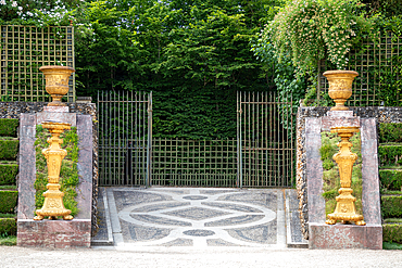 Formal garden entrance with decorative stone pavement, iron gates, sculpted golden urns and candelabras, and green trellis with greenery at Palace of Versailles, Paris.
