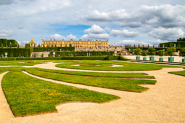 Versailles gardens with geometric lawns and gravel paths, leading to the palace and Orangerie under a partly cloudy sky at Palace of Versailles, Paris.
