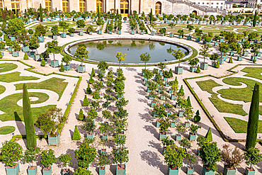 Aerial view of a formal garden with a circular pond, potted trees, topiary, and gravel paths, adjacent to a large building at Palace of Versailles, Paris.