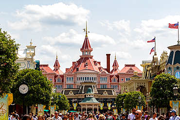 A crowd of people on a street lined with themed buildings, including a pink hotel with red roofs, under a partly cloudy sky at Disneyland Paris, France.