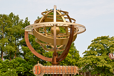 The image shows the Discoveryland sign at Disneyland Paris, featuring a golden armillary sphere against a backdrop of green trees and a pale sky at Disneyland Paris, France.