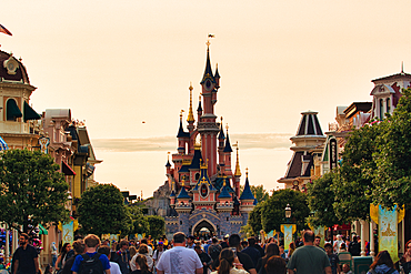 Crowd on a street leads towards a pink and blue castle under a warm, hazy sky at a theme park.