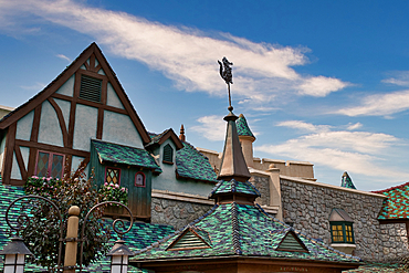 Low-angle view of various buildings with unique architecture, stone walls, and green-blue tiled roofs, under a blue sky with scattered clouds at Disneyland Paris, France.