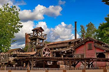 A wooden mining structure dominates the frame with weathered textures, under a blue sky with puffy clouds and framed by green trees at Disneyland Paris, France.