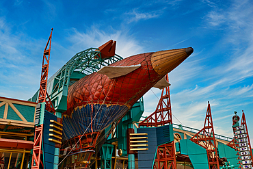 A large airship structure, primarily red, blue, and gold, sits amidst teal and orange metal framework under a bright blue sky with wispy clouds at Disneyland Paris, France.
