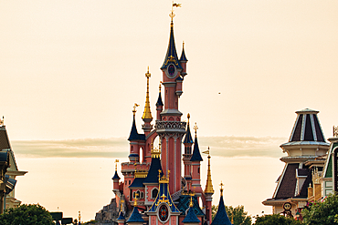Centered view of a pink fairytale castle with blue turrets, gold accents, and other buildings visible in the foreground against a pale sky at Disneyland Paris, France.