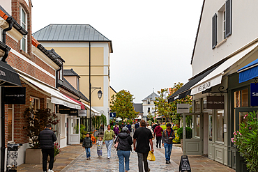 Outdoor shopping area with brick and beige buildings, awnings, and pedestrians walking on a stone-paved street under an overcast sky at La Vallee Village, France.