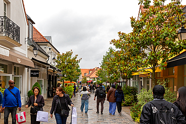 Outdoor scene of people walking on a cobblestone street lined with shops and trees under a cloudy sky at La Vallee Village, France.