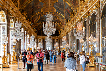 The Hall of Mirrors at Versailles is filled with visitors. Chandeliers hang from the ceiling, reflecting in the mirrored walls.