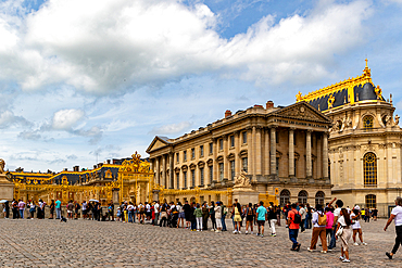 Exterior view of the Palace of Versailles with a crowd of tourists in the foreground under a cloudy sky.