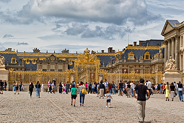 A crowd of people walks across a stone courtyard in front of a large building with golden gates and intricate architectural details.