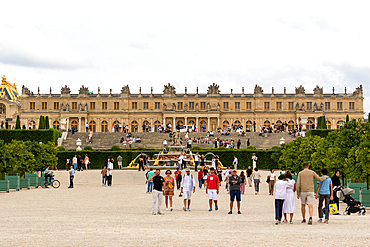 Palace of Versailles exterior, featuring a large crowd of people, gardens and a fountain on a cloudy day.