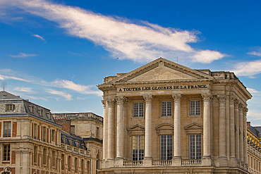 The image features a neoclassical building with columns and a pediment, part of a larger complex under a blue sky with clouds at Versailles Gardens, Paris.