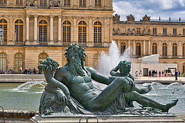 Bronze Neptune statue in a fountain at Versailles. The Palace of Versailles is in the background under a cloudy sky.