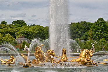Golden statues of horses, figures, and a chariot in a fountain spraying water, with trees and buildings in the background under a cloudy sky at Versailles Gardens, Paris.