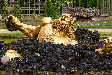 A golden statue of a man is partially submerged in black rocks, in water, in front of a manicured garden at Versailles Gardens, Paris.