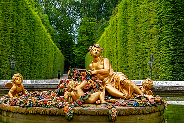 Gold sculptures on flower-covered fountain in a garden with green hedges at Versailles Gardens, Paris.