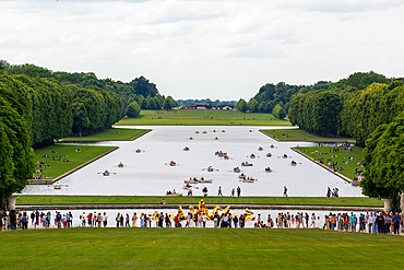 Wide angle shot of a large pond with rowboats, flanked by manicured grass and trees, viewed from a grassy hill with a crowd in front at Versailles Gardens, Paris.