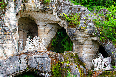 Three grottoes containing white statues carved into rock are covered with moss and greenery under natural light at Versailles Gardens, Paris.