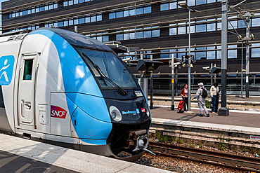 A blue and white SNCF train at a station, with people waiting on the platform. Modern building in the background.