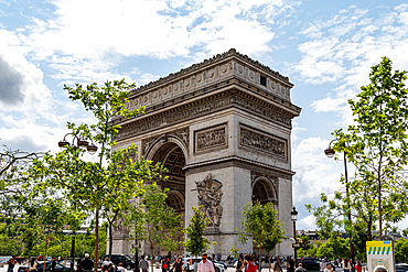 The Arc de Triomphe in Paris is centered, framed by trees and street lamps, with people visible in the foreground under a partly cloudy sky.