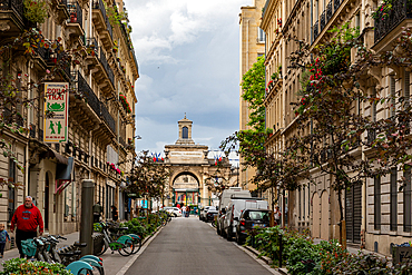 Parisian street view featuring architecture, parked vehicles, pedestrians, and the Hôpital Lariboisière in the background under a cloudy sky.