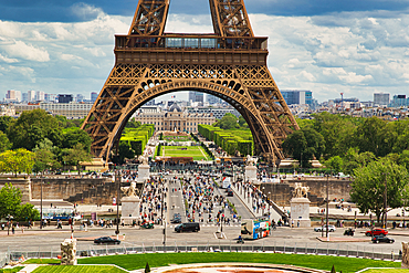 View through the Eiffel Tower archway shows a cityscape with a garden, buildings, many people, and traffic on a cloudy day.