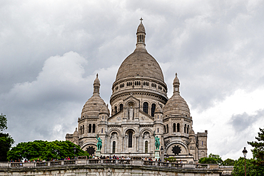 A low-angle view of the Sacré-Cœur Basilica in Paris, featuring its domes, architecture, and surrounding greenery under a cloudy sky.