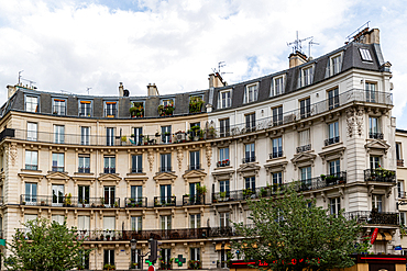 Building facade with multiple windows and balconies, featuring architectural details and plants, under an overcast sky.