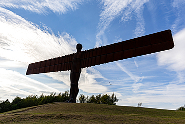 A silhouette of the Angel of the North sculpture against a blue sky with streaky clouds, atop a grassy hill.