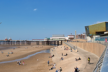 Beach scene with pier, modern building, and many people on a sunny day under a blue sky.