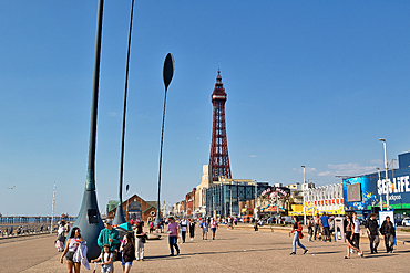 Blackpool promenade featuring the Blackpool Tower, various buildings, and people walking under a clear blue sky.