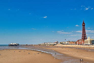 Wide shot of Blackpool beach, pier, and tower. People scattered across the sand under a bright blue sky.