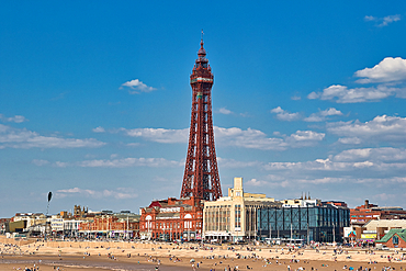 Wide shot: Blackpool Tower dominates the skyline, with buildings and a crowded beach below, under a blue sky with scattered clouds.