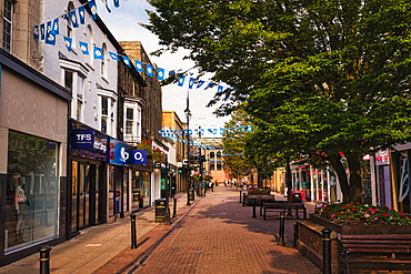 A pedestrian street is lined with shops, benches, and large trees. Blue and white flags are strung above the street in Harrogate, UK.