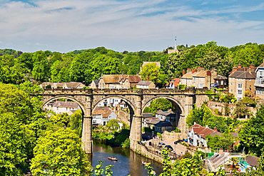 Scenic view of a stone viaduct spanning a river, surrounded by lush greenery and houses under a partly cloudy sky in Knaresborough, UK.