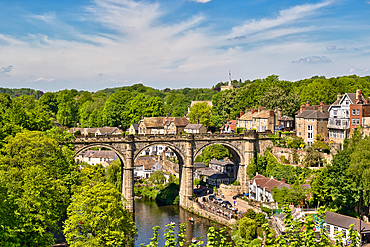 Scenic view of a stone viaduct with buildings atop, spanning a river in a lush, green landscape under a blue sky with scattered clouds in Knaresborough, UK.