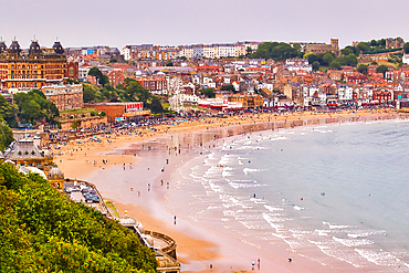A panoramic view of a crowded beach with a town and a ferris wheel overlooking the water in Scarborough, UK.