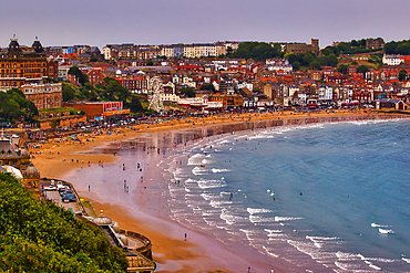 A wide view of a bustling beach town with a sandy beach, blue ocean, and densely packed buildings in Scarborough, UK.