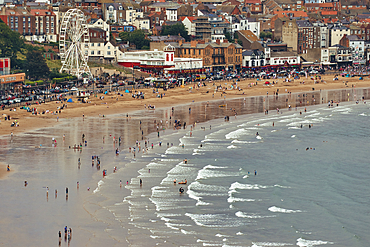 A bustling beach scene with many people, buildings, a Ferris wheel, and the ocean with waves in Scarborough, UK.