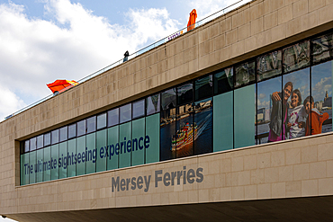 A modern building facade displays large windows with promotional graphics, featuring the text 'Mersey Ferries' and images of a ferry and a family in Liverpool, UK.