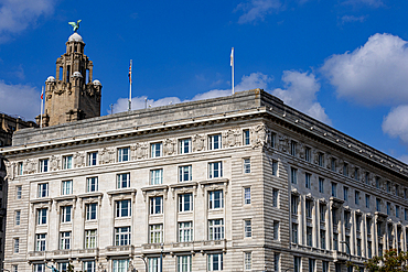 A grand stone building with many windows and decorative elements, under a blue sky with clouds. A tower with a bird sculpture stands to the left in Liverpool, UK.